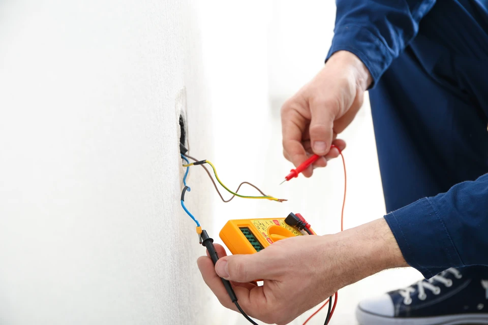 Électricien en tenue bleue effectuant des mesures électriques sur installation murale avec multimètre numérique orange