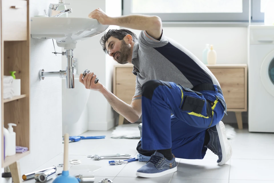 Plombier professionnel en tenue de travail bleue effectuant une réparation sous un évier de cuisine moderne