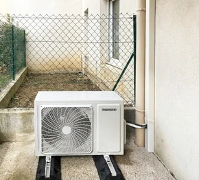 Unité extérieure de pompe à chaleur air/air marque Thermor, installée en extérieur contre un mur blanc, posée sur plots anti-vibratoires