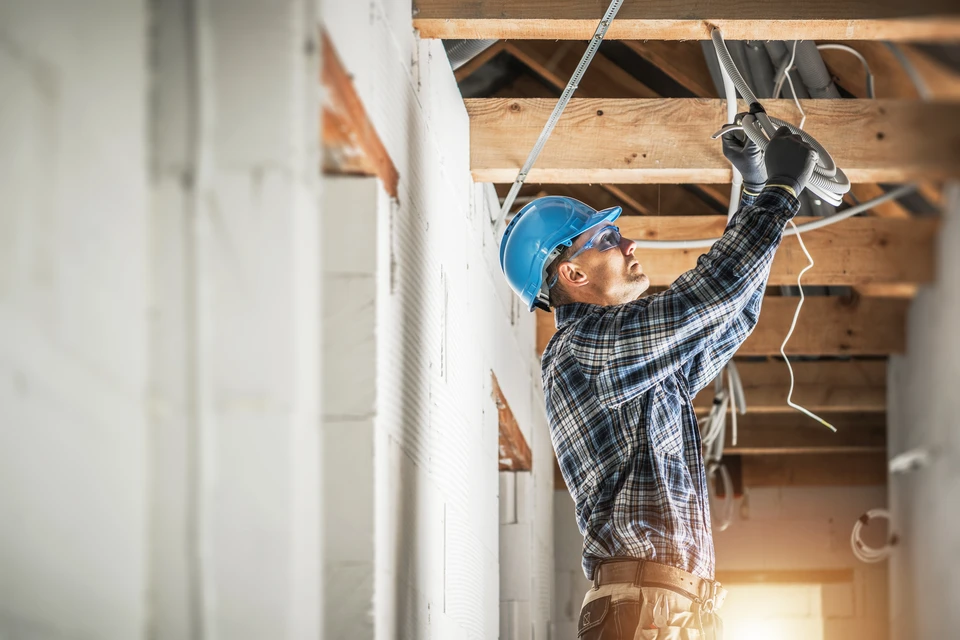 Photographie d'un électricien en tenue professionnelle, équipé d'outils, effectuant l'installation ou la maintenance d'un tableau électrique dans un intérieur résidentiel ou professionnel. L'électricien doit être photographié en train de travailler avec concentration, démontrant la précision et le professionnalisme. Le tableau électrique doit être clairement visible avec ses disjoncteurs organisés et correctement installés. L'environnement doit être un intérieur réel (mur, peinture) sans éléments de fantaisie. L'éclairage naturel ou professionnel doit valoriser le côté métier et la confiance.